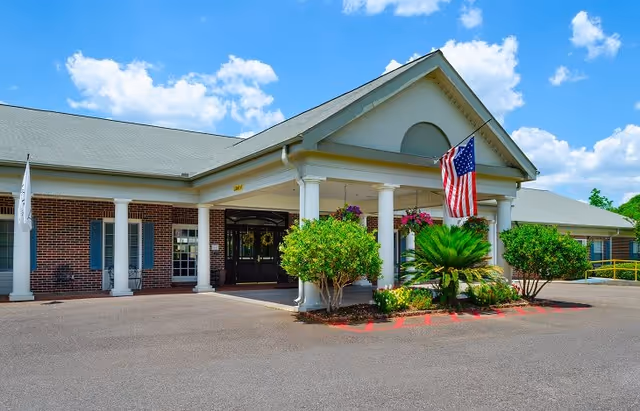 Front exterior view of a single-story brick building with white columns and a covered entrance. There are green shrubs and plants near the entrance, and an American flag is displayed on a pole attached to the building. The sky is blue with scattered clouds.