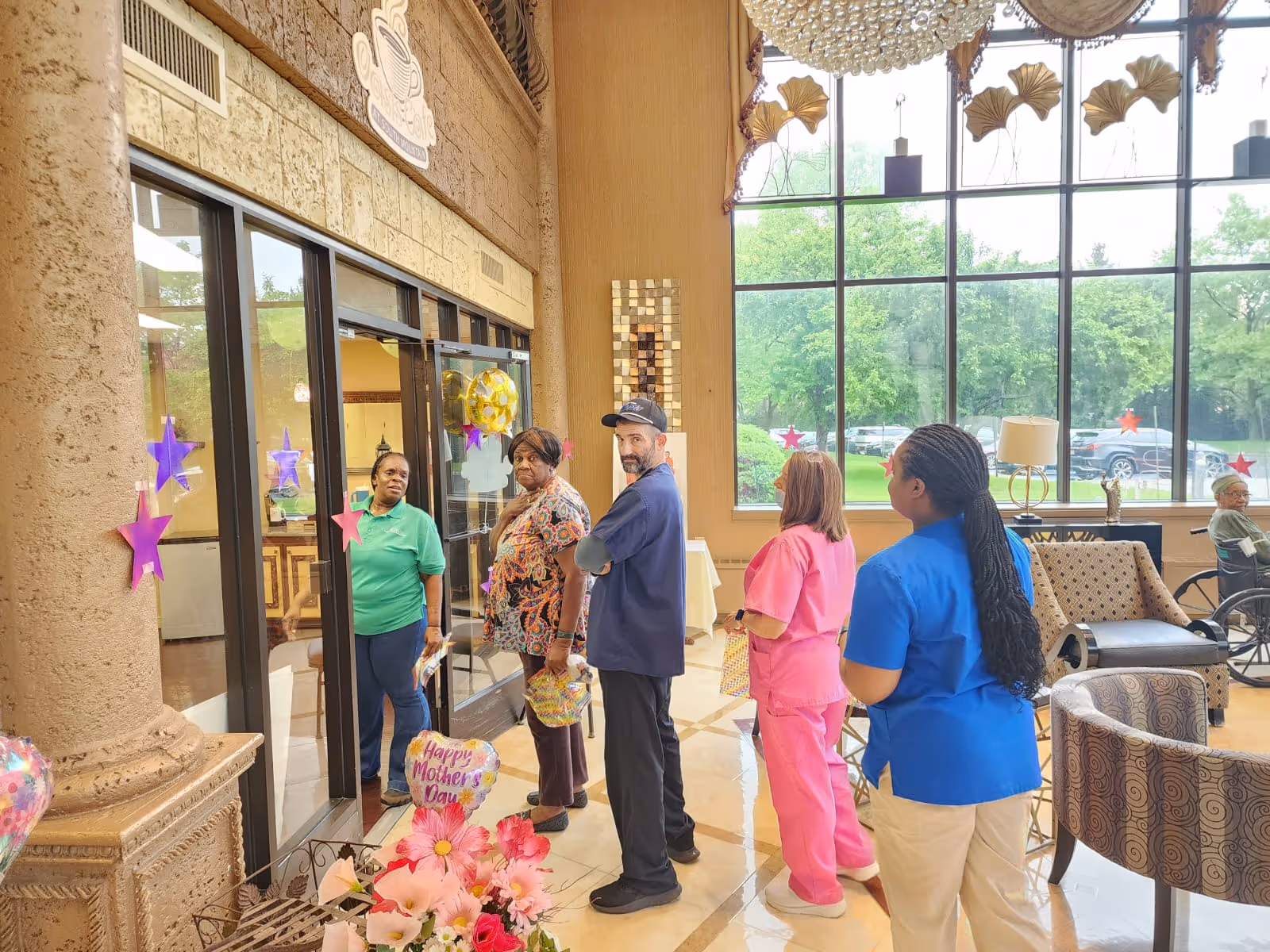 A group of five people, including healthcare workers in scrubs and casual clothing, standing in a bright lobby area decorated with star-shaped stickers on the glass doors and windows. There are chairs and a wheelchair visible near large windows showing a green outdoor area with parked cars. A bouquet of flowers with a 'Happy Mother's Day' balloon is in the foreground.