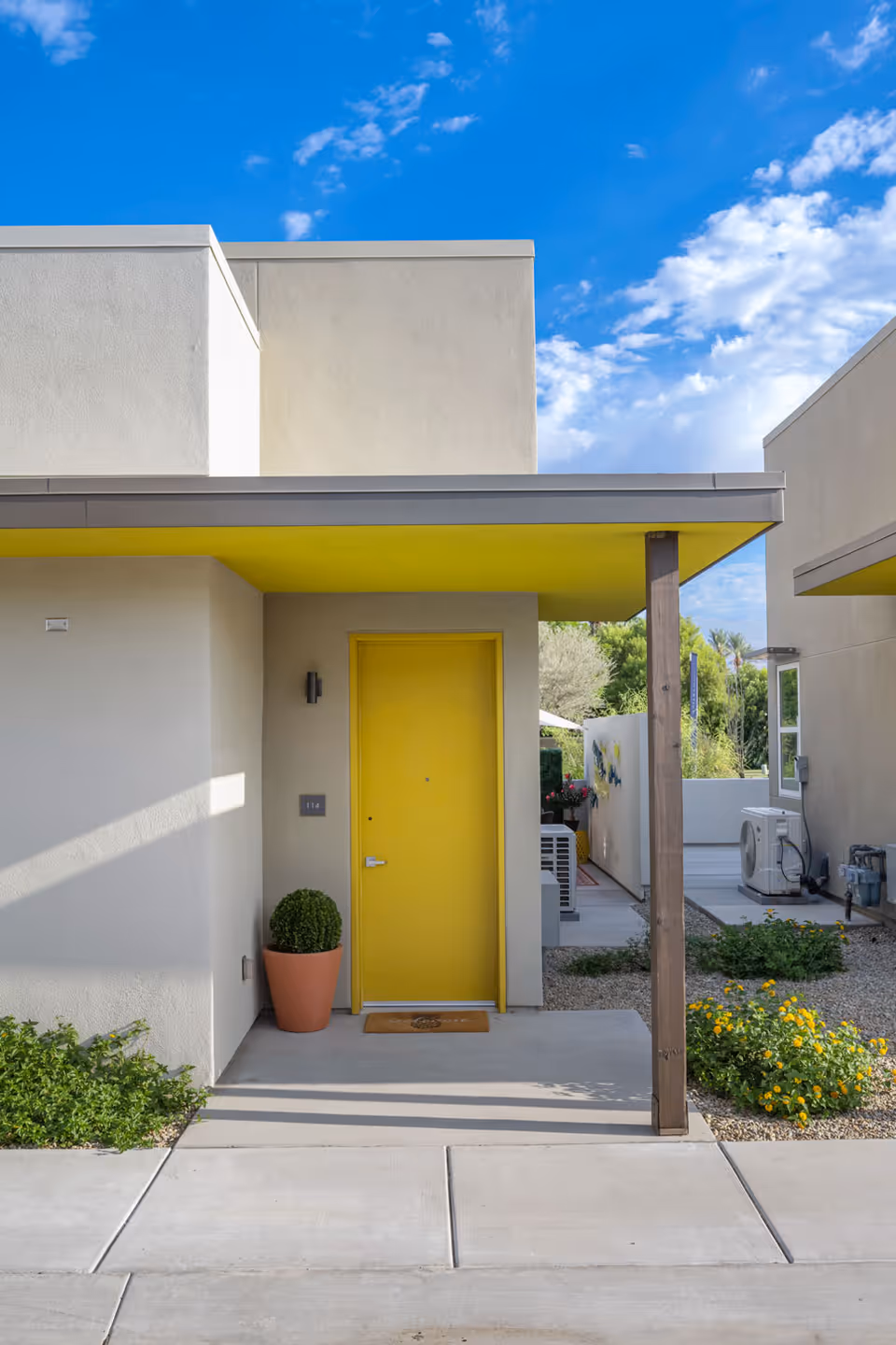 Modern building entrance with a bright yellow door and matching canopy, a potted plant, and a walkway under a blue sky.