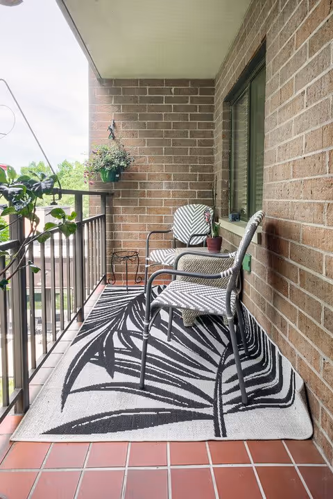 A small balcony with brick walls and a tiled floor, featuring two patterned chairs, a small table with a potted plant, a hanging plant on the railing, and a black and white rug with a leaf design.