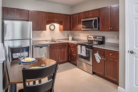 A modern kitchen with dark wooden cabinets, stainless steel appliances including a refrigerator, dishwasher, microwave, and stove. There is a small wooden dining table with two chairs, set with plates, napkins, and wine glasses. A clock is mounted on the wall above the sink.