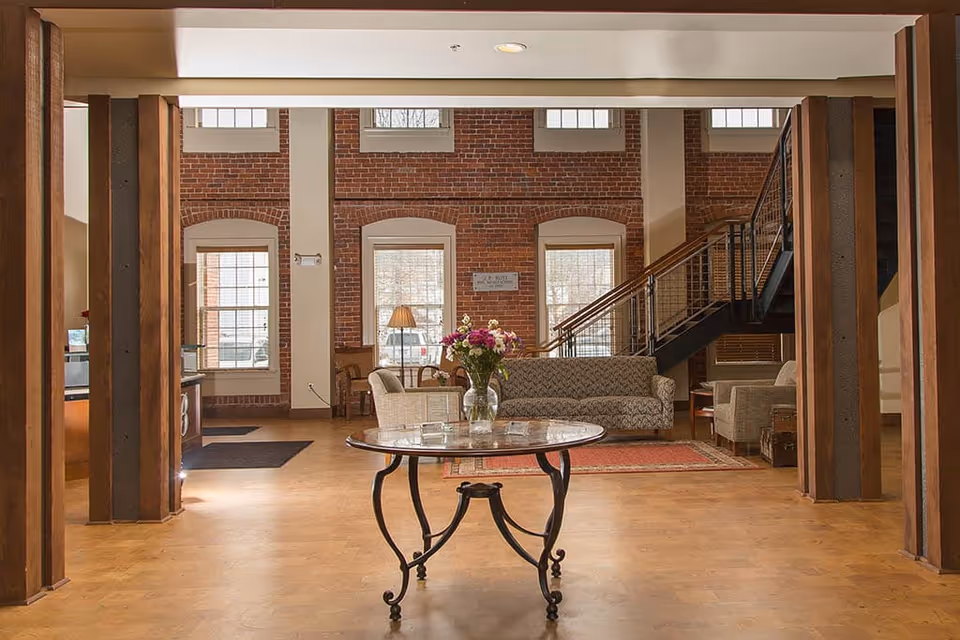 A spacious interior common area with wooden floors and exposed brick walls. The room features a round glass table with a vase of flowers in the center, a patterned sofa, armchairs, a floor lamp, and a staircase with metal railings leading to an upper level. Large windows allow natural light to fill the space.