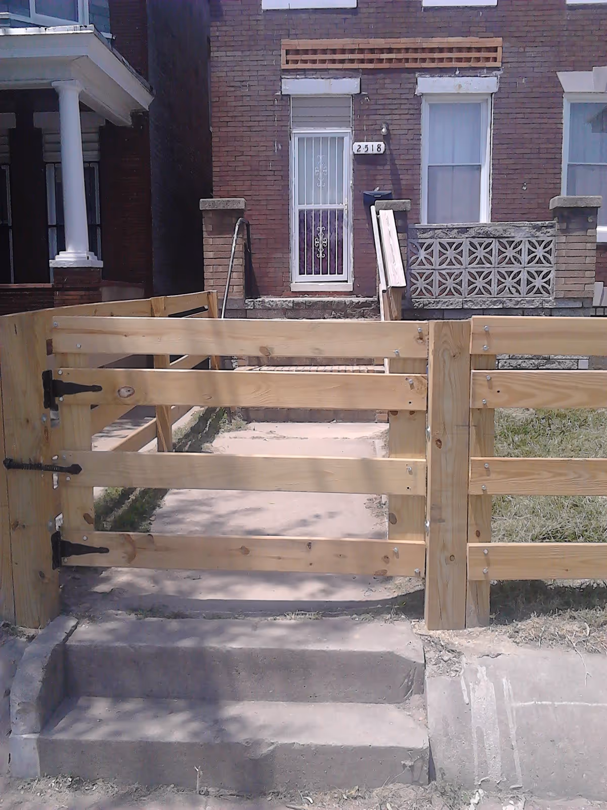 Front exterior view of a brick residential building with a white metal security door and two windows. There is a wooden fence and gate in front of the building, with concrete steps leading up to the gate and the entrance. The building number 2518 is displayed next to the door.