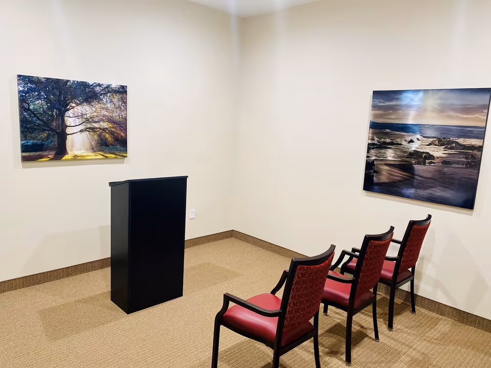Small meeting room with a black podium facing three red upholstered chairs and two landscape paintings on cream walls.