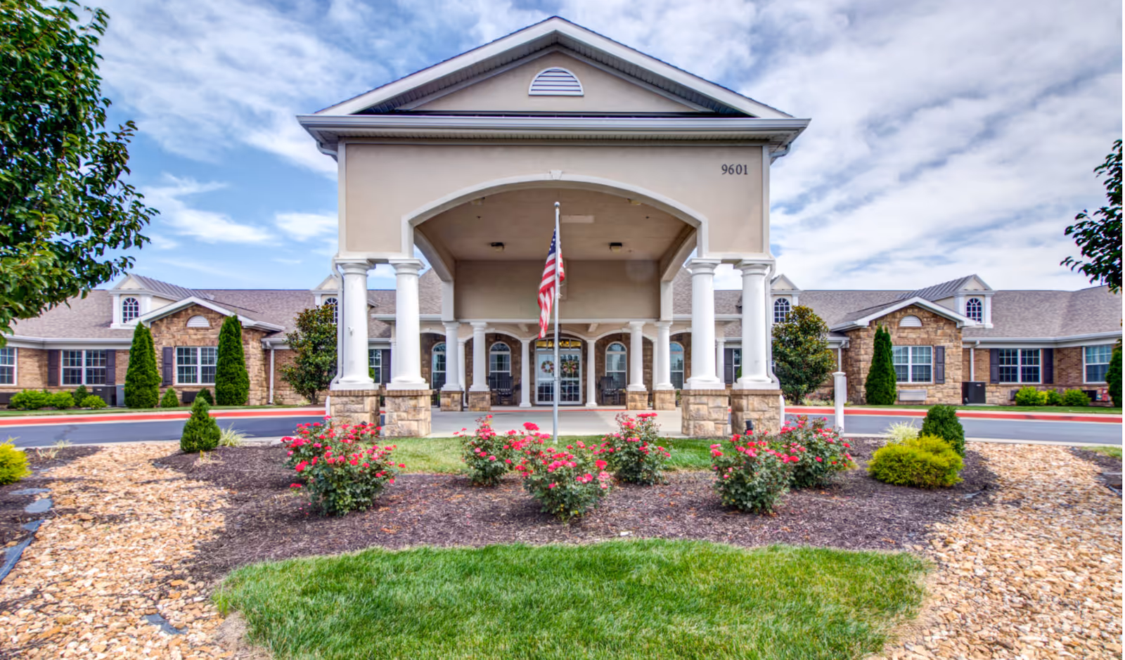 Front exterior view of Addington Place of Shoal Creek, featuring a covered entrance with white columns, an American flag in the center, landscaped flower beds with red flowers, and a clear blue sky with some clouds.