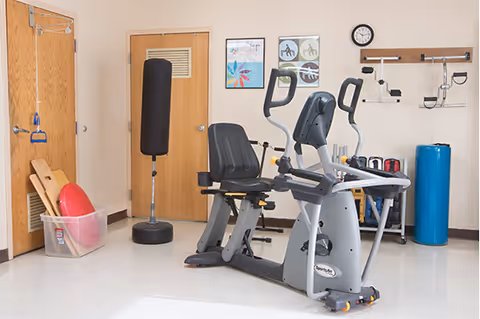 A small exercise room with a recumbent stationary bike, a standing punching bag, a plastic bin containing exercise equipment, a blue foam roller, and various exercise tools mounted on the wall. The room has beige walls, a clock, and two wooden doors.