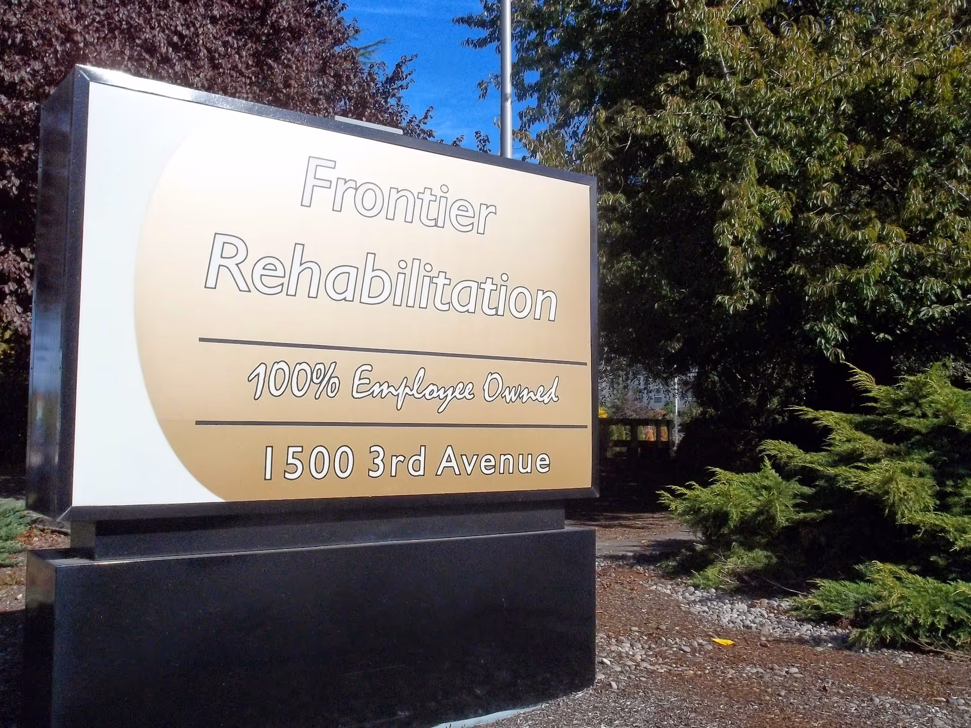 Outdoor sign reading 'Frontier Rehabilitation 100% Employee Owned 1500 3rd Avenue' in front of trees and shrubs.