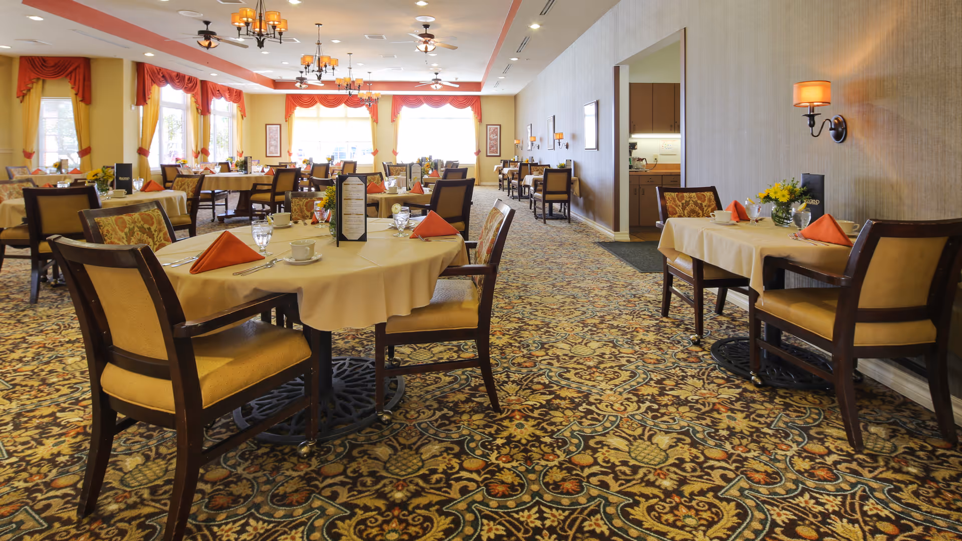 A spacious dining room with multiple tables covered in beige tablecloths, each set with orange folded napkins, glassware, and floral centerpieces. The room features patterned carpet, large windows with red and yellow curtains, wall sconces, and chandeliers providing warm lighting.