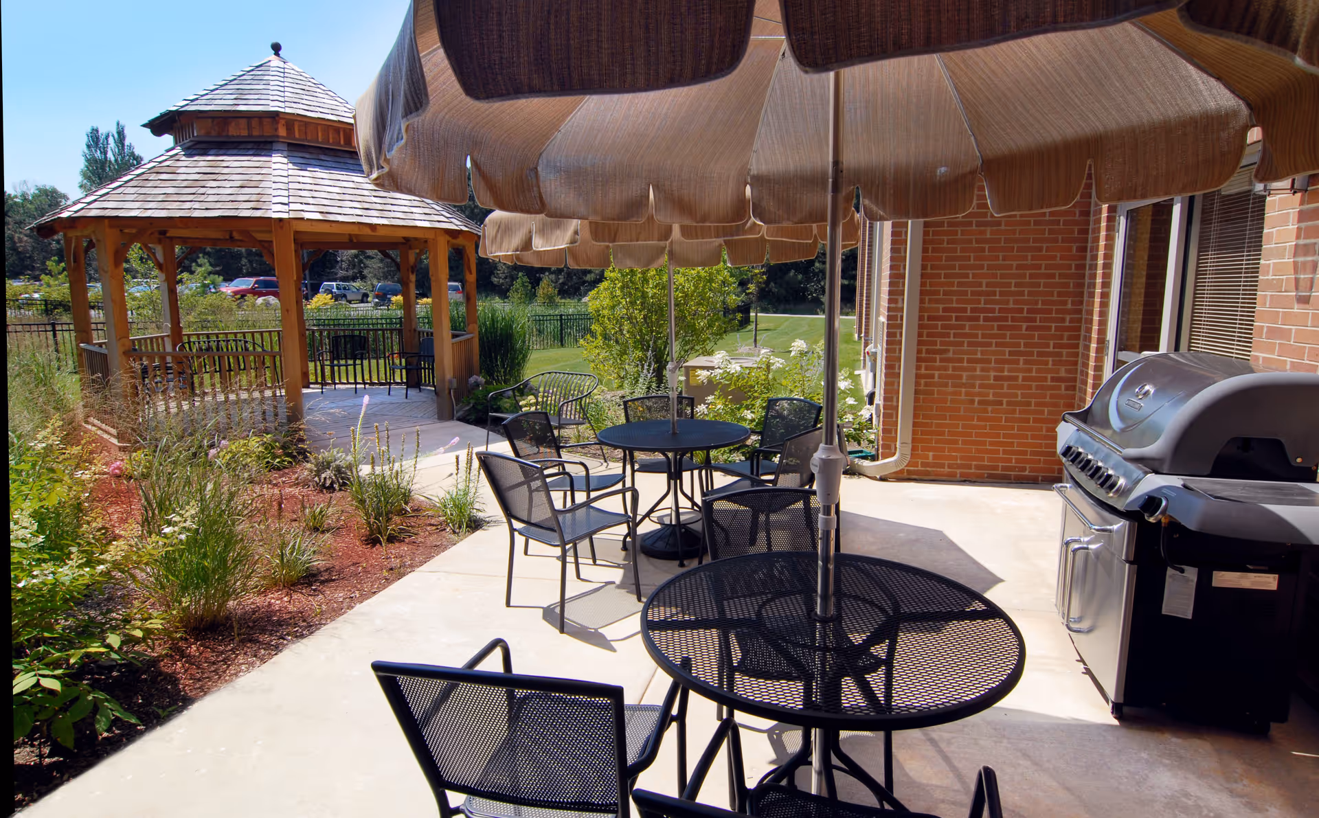 Outdoor patio with round metal tables and umbrellas, chairs, a wooden gazebo, and a grill beside a brick building.