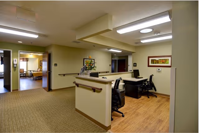 Interior view of a senior living facility reception or nurse station area with two office chairs and desks. The area has beige walls, wood flooring in the workstation area, and carpet in the hallway. There are handrails along the walls and framed artwork hanging. An open door leads to a room with a bed and chairs visible.