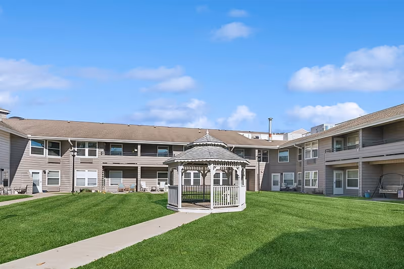 A courtyard area of a senior living facility with a green lawn, a white gazebo in the center, and a two-story building surrounding the courtyard under a blue sky with some clouds.
