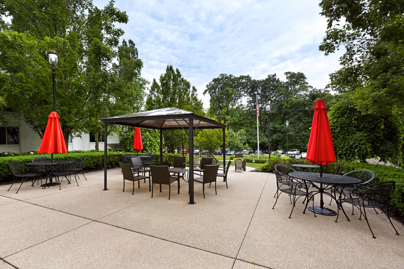 Outdoor patio area with several black metal tables and chairs, each table shaded by a bright red umbrella. A gazebo with additional seating is in the center. The area is surrounded by green trees and bushes, with a parking lot and American flag visible in the background under a partly cloudy sky.