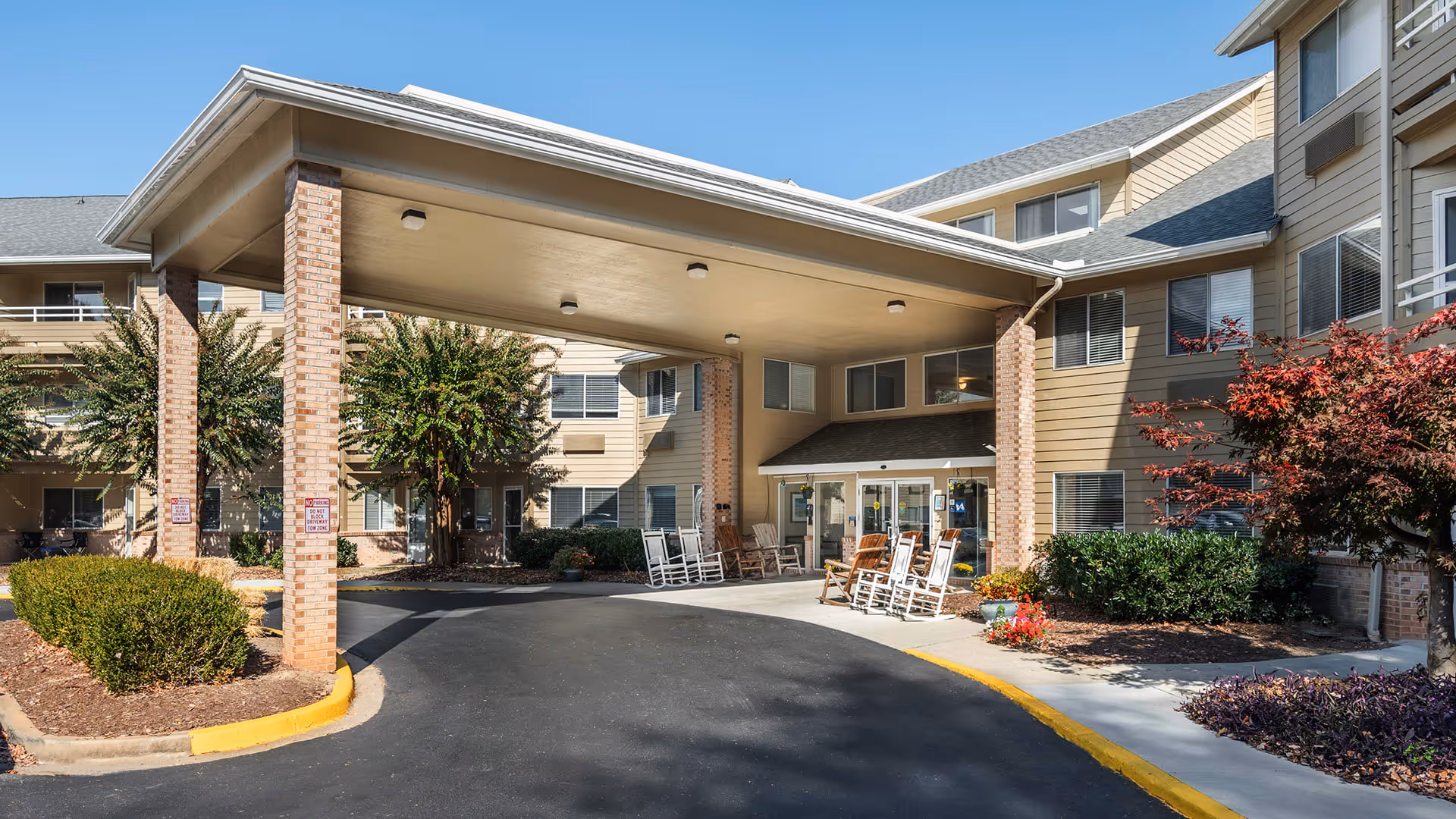 Entrance of a senior living facility with a covered drop-off area supported by brick columns. Several rocking chairs are placed near the glass doors of the building. The building is three stories tall with beige siding and multiple windows. There are landscaped bushes and trees around the entrance under a clear blue sky.