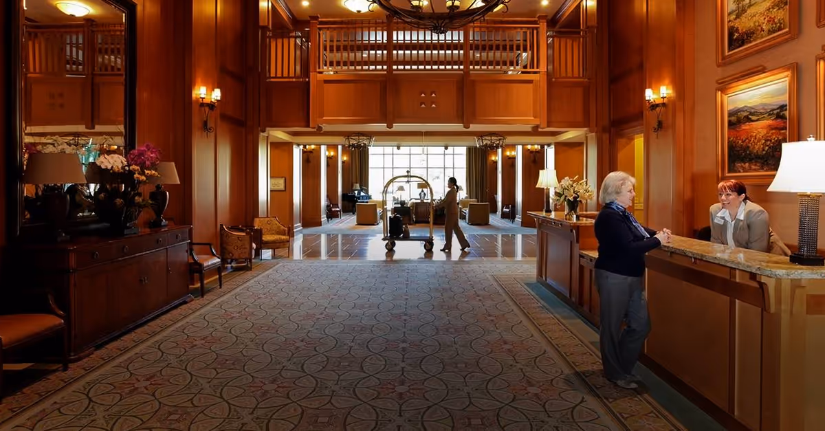 A spacious and warmly lit senior living facility lobby with wooden paneling and a patterned carpet. An elderly woman is speaking with a receptionist at a wooden front desk on the right. In the background, a staff member is pushing a luggage cart towards a seating area with large windows letting in natural light. The lobby is decorated with flowers, lamps, and framed paintings on the walls.