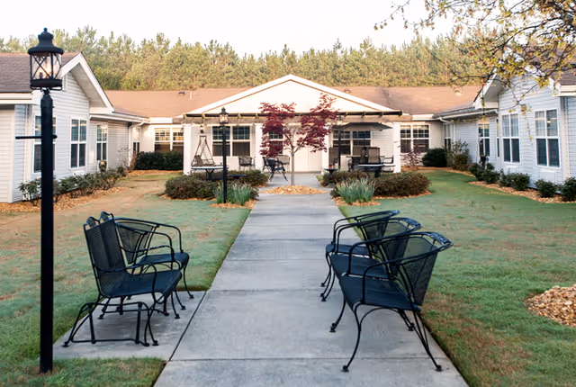 Outdoor courtyard area of a senior living facility with a concrete pathway leading to a building entrance. The pathway is flanked by black metal benches on both sides and a black lamp post on the left. The building is single-story with white siding and multiple windows, surrounded by grass, shrubs, and small trees.