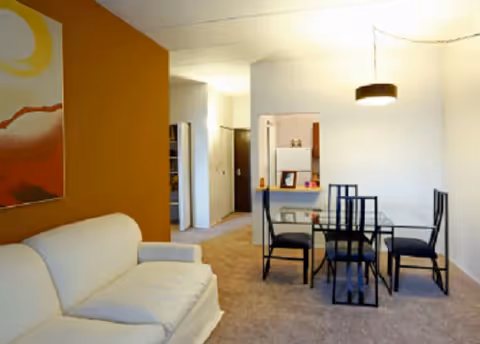 Open-plan living and dining area with a white sofa, glass dining table with black chairs, and a warm-toned accent wall.