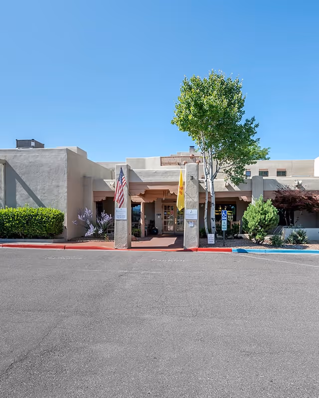 Front exterior view of Brookdale Santa Fe facility with a clear blue sky, two flags on pillars at the entrance, and surrounding greenery including trees and bushes.
