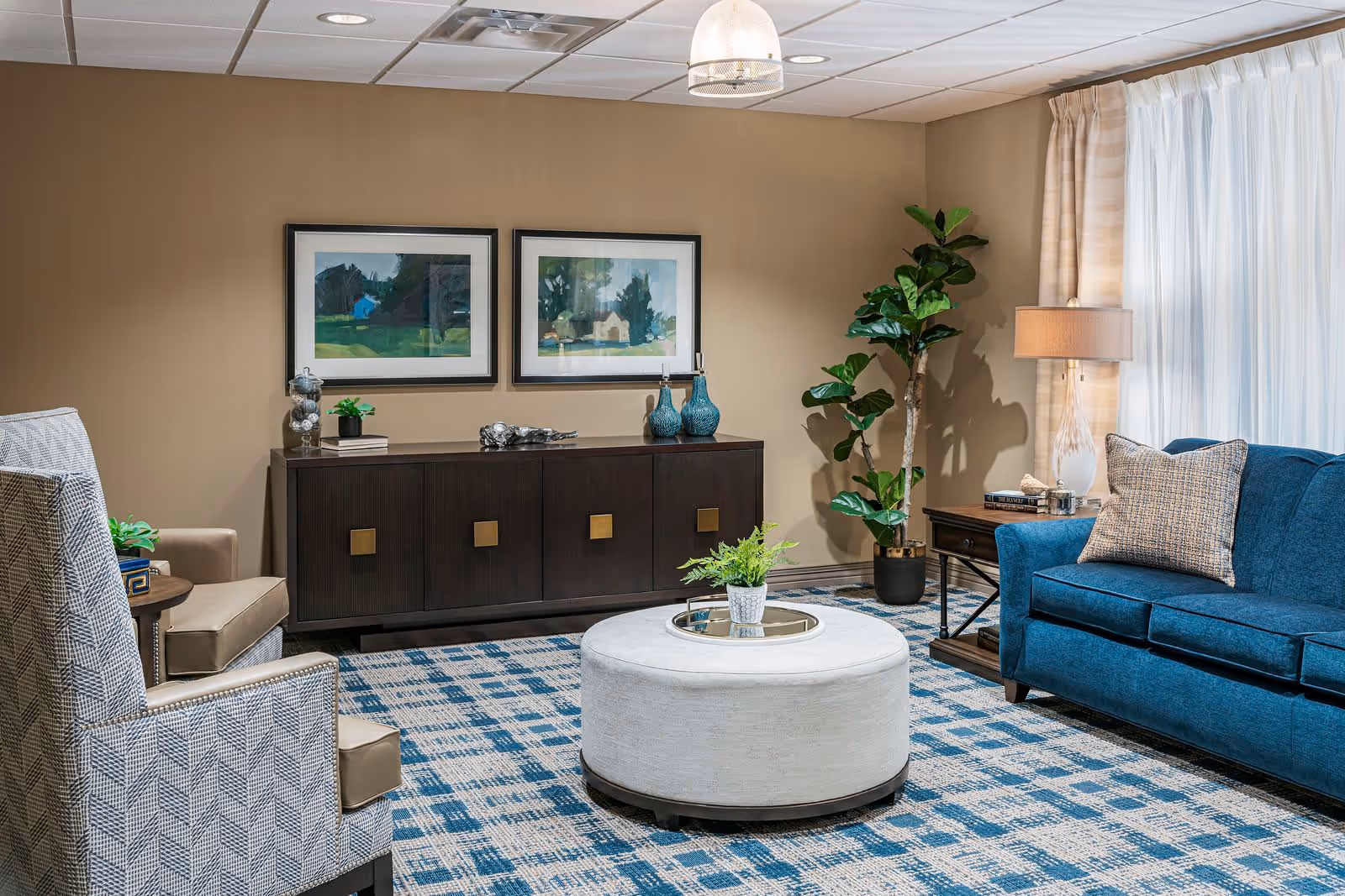 A cozy living room with a blue sofa, a patterned armchair, a round white ottoman with a small plant on top, a dark wooden sideboard with decorative items, two framed landscape paintings on the wall, a tall green plant in a pot, a side table with a lamp and books, and large windows with sheer white curtains.