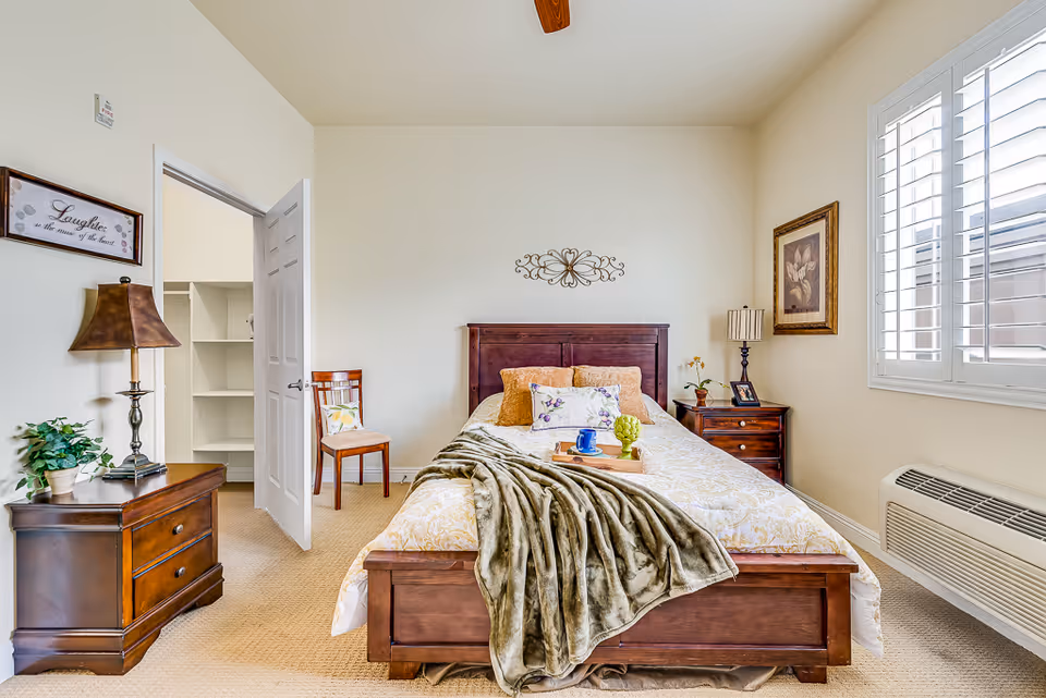A bright and cozy bedroom with a wooden bed frame, beige bedding, and decorative pillows. A green throw blanket is draped over the bed, which has a tray with a blue cup and a small green plant. There are two wooden nightstands on either side of the bed, each with a lamp and decorative items. A wooden chair with a cushion is placed near an open door leading to a walk-in closet with shelves. The room has beige carpet, white walls, a ceiling fan, and a window with white shutters letting in natural light.