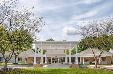 Front exterior view of Lighthouse Senior Living at Ellicott City showing a single-story building with a covered entrance, surrounded by trees and a clear sky with some clouds.