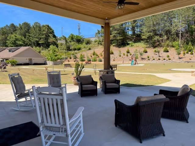Covered outdoor patio area with several chairs including white rocking chairs and dark wicker chairs arranged on a concrete floor. Beyond the patio is a grassy area with a playground, benches, and a backdrop of trees and a hill under a clear blue sky.