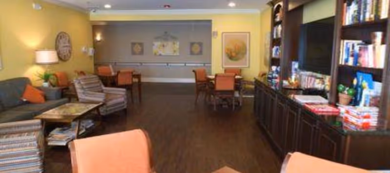 A communal lounge with sofas, armchairs, tables, and a bookshelf/TV against a yellow wall.