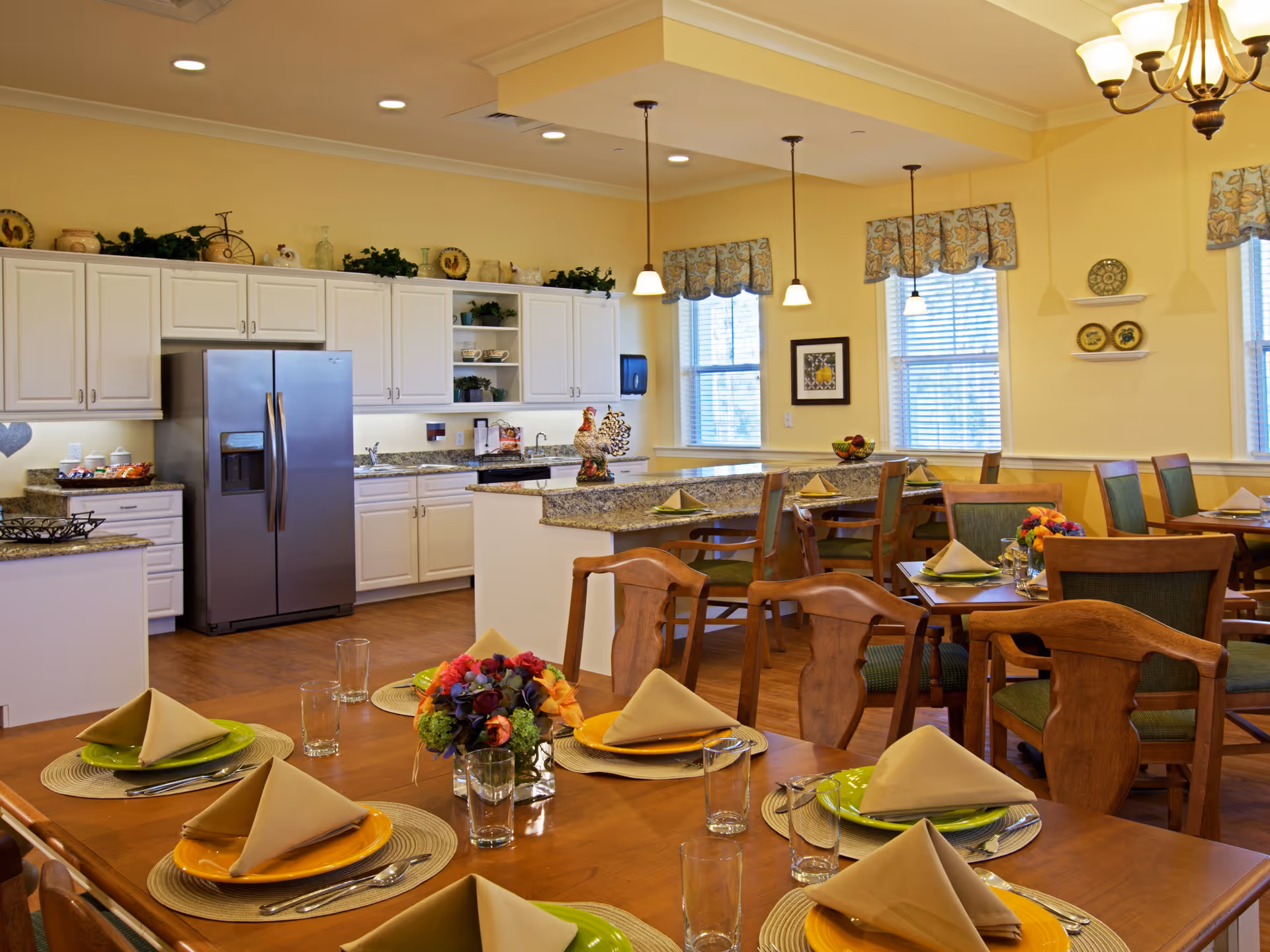 A bright dining area with wooden tables set with colorful plates, beige folded napkins, glasses, and silverware. The room features a kitchen area with white cabinets, a stainless steel refrigerator, granite countertops, and decorative items on top of the cabinets. There are large windows with patterned valances allowing natural light to fill the space, and pendant lights hanging from the ceiling.