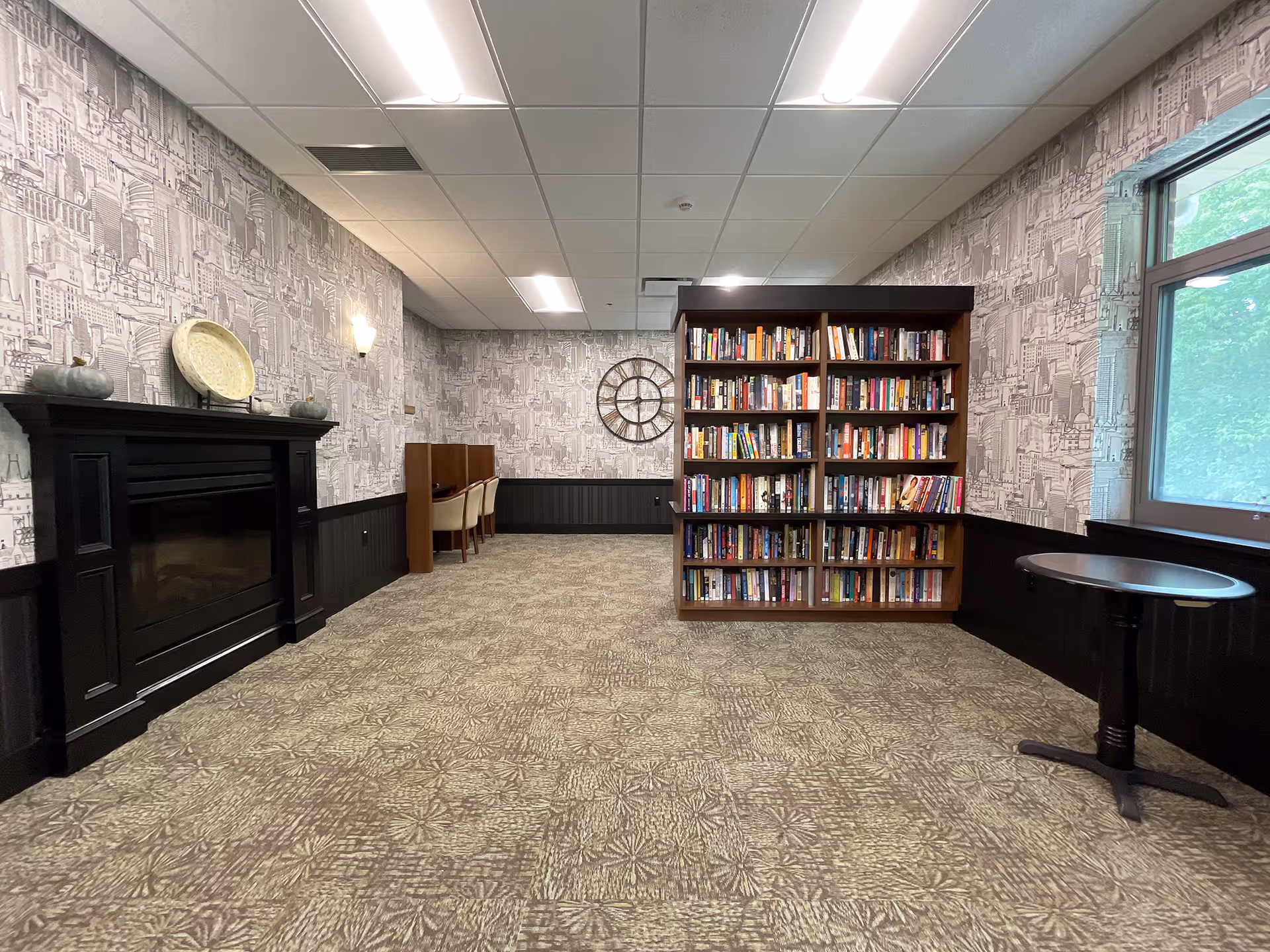 A quiet room with patterned carpet and wallpaper featuring cityscape designs. There is a black fireplace on the left wall with decorative items on top, a wooden bookshelf filled with books in the center, and a small round table near a large window on the right. Two chairs and a small desk are positioned against the back wall under a large decorative clock.