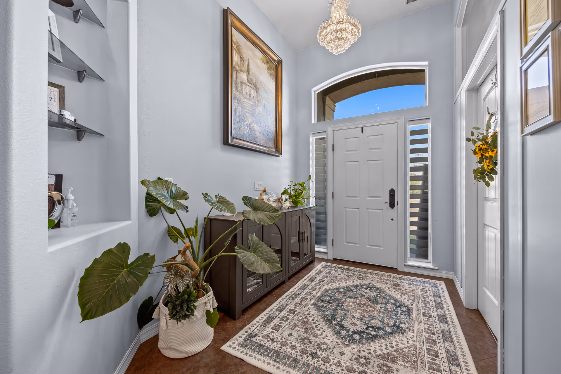 Bright foyer with a white front door, patterned rug, console table, potted plants, and a chandelier.
