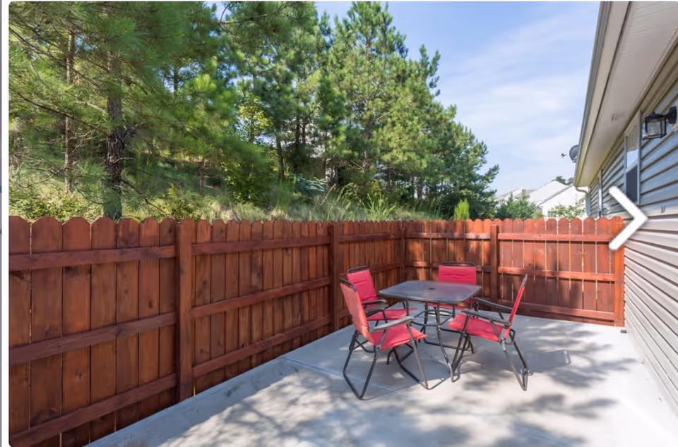 Outdoor patio area with a concrete floor, surrounded by a wooden fence. There is a square table with four red cushioned chairs arranged around it. Trees and greenery are visible beyond the fence under a partly cloudy sky.