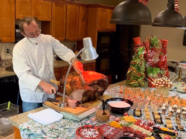 A chef wearing a white coat and face mask is carving a large piece of roasted meat under a heat lamp in a kitchen setting. The countertop is covered with a festive tablecloth and is filled with an assortment of appetizers, desserts, and decorative food displays, including a Christmas tree-shaped arrangement made of green and red items.