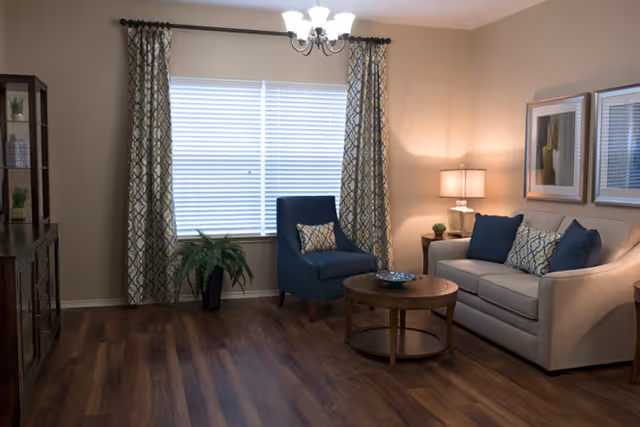 A cozy living room with a beige sofa adorned with blue and patterned cushions, a blue armchair with a matching cushion, a round wooden coffee table, a side table with a lamp, two framed artworks on the wall, patterned curtains on a window, a potted plant, and a wooden shelving unit with decorative items.