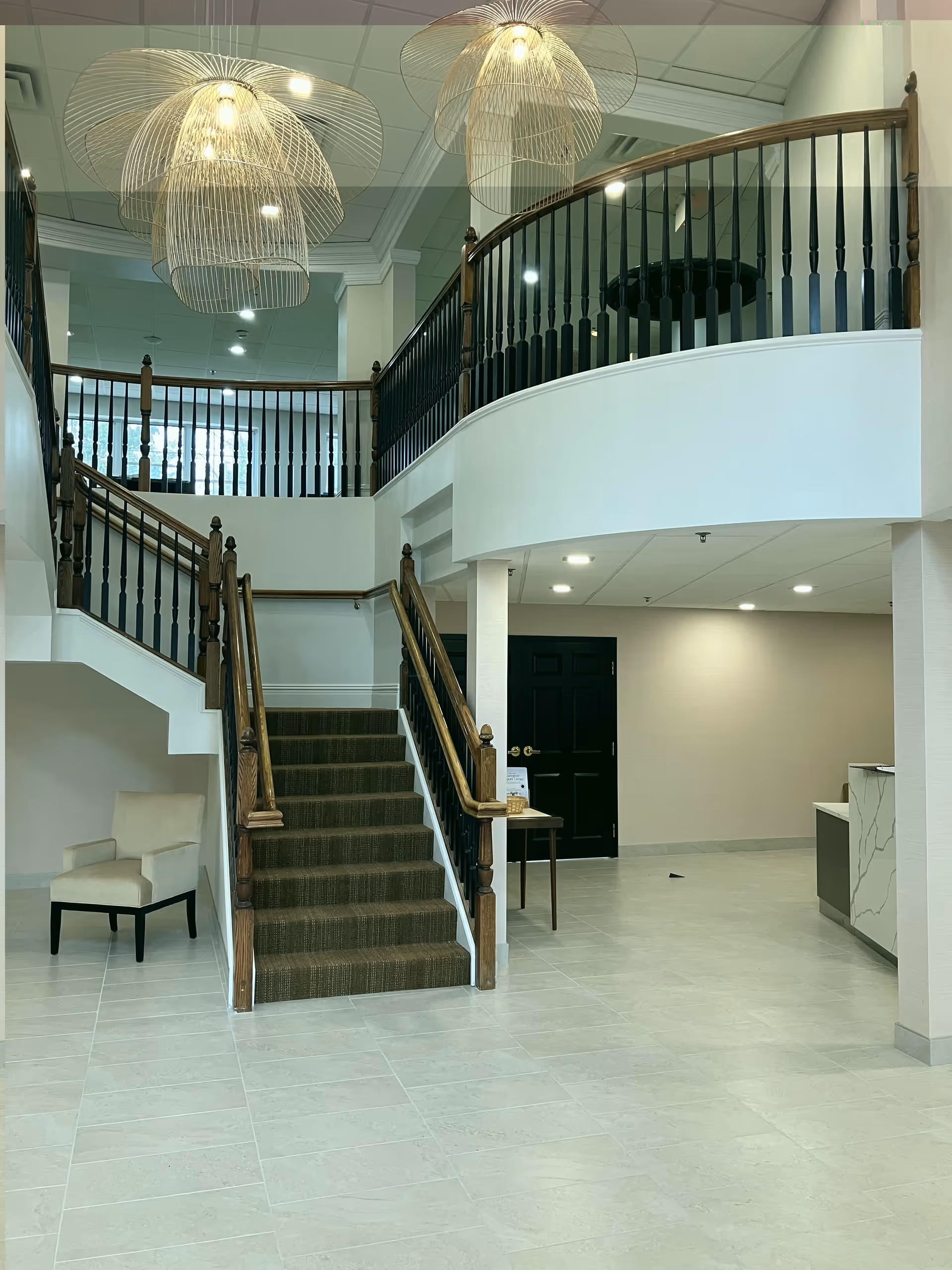 Interior view of a senior living facility lobby with a staircase leading to an upper floor. The area features modern pendant lights, a beige armchair, a small table with a basket, and a reception desk. The floor is tiled, and the walls are painted in neutral tones.
