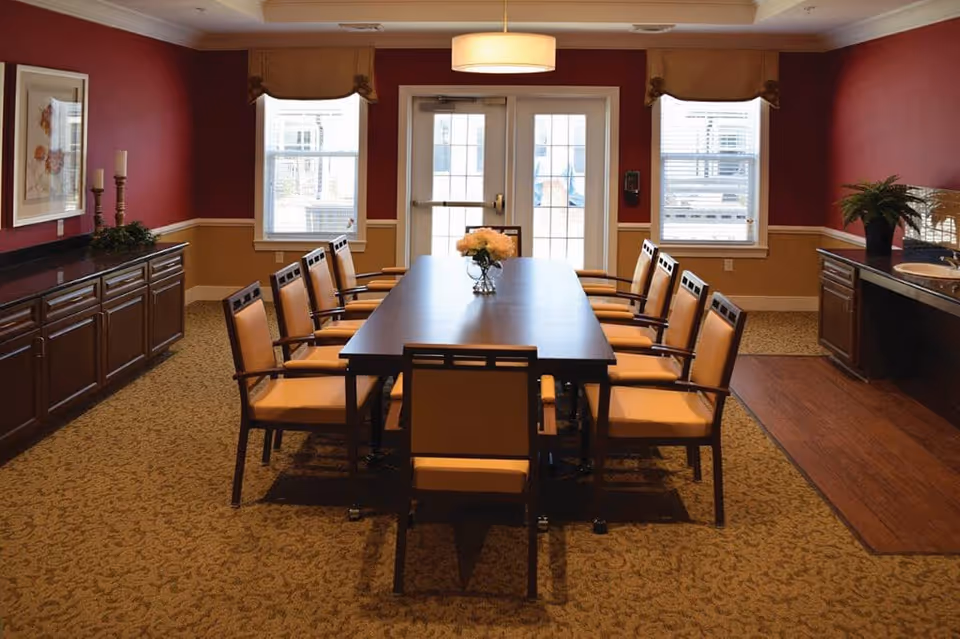 A dining room with a long rectangular table surrounded by ten chairs with tan cushions. The walls are painted red with a beige wainscot, and there are two windows and a glass door letting in natural light. The room features a sideboard with decorative candles and a plant, and a small sink area with a potted plant. A ceiling light fixture hangs above the table.