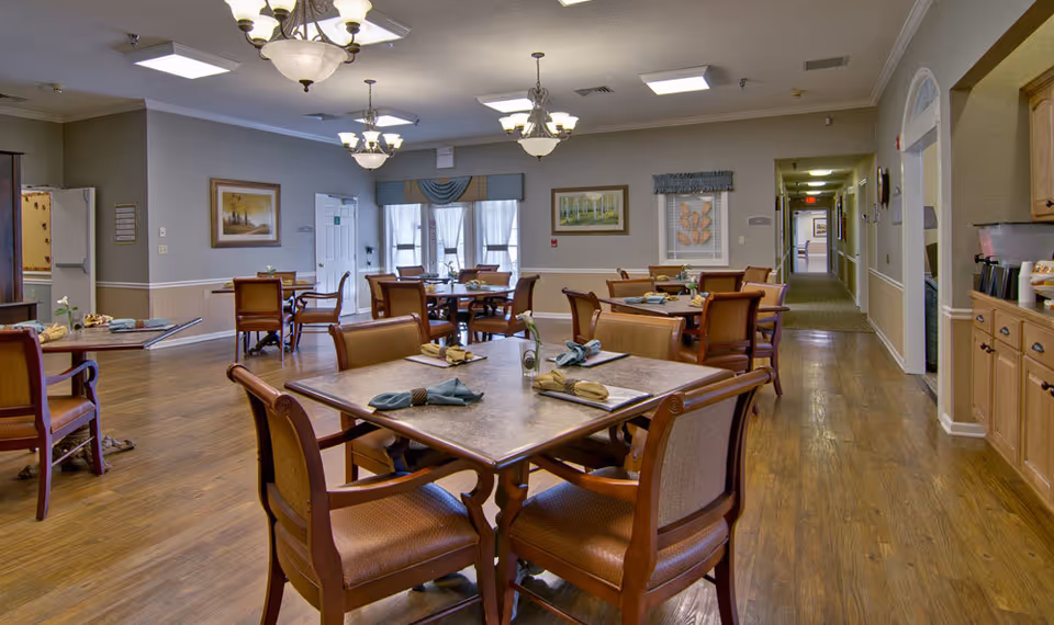 A senior living facility dining room with multiple wooden tables and chairs arranged neatly. Each table is set with napkins and menus. The room has wooden flooring, soft lighting from ceiling chandeliers, framed artwork on the walls, and large windows with curtains letting in natural light. A hallway is visible in the background.