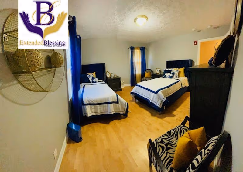A bedroom with two single beds dressed in white and navy blue bedding. The room has light wood flooring, two windows with blue curtains, a nightstand between the beds, a black dresser, and a zebra-patterned chair with yellow cushions. A decorative wall piece is visible on the left wall. The logo of Extended Blessing Personal Care Home is overlaid in the top left corner.