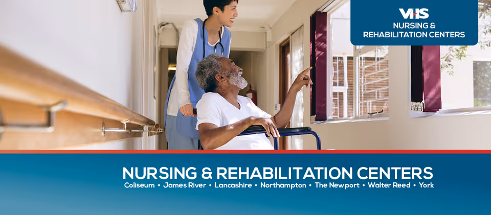 A healthcare worker in a light blue uniform is pushing an elderly man in a wheelchair down a hallway with handrails. The elderly man is pointing towards something outside a window on the right side. The setting appears to be inside a nursing and rehabilitation center.
