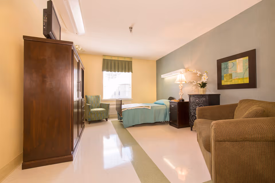 A well-lit nursing facility room with a single bed covered in a teal blanket, a wooden nightstand with a lamp and a flower vase, a brown armchair, a green striped armchair near a window with a green valance, and a large wooden wardrobe with a TV on top.