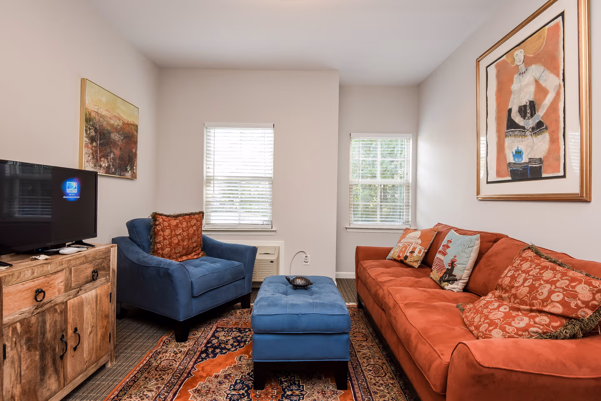 Cozy living room with a red sofa, blue armchair and ottoman, wooden TV console, patterned rug, and framed artwork.