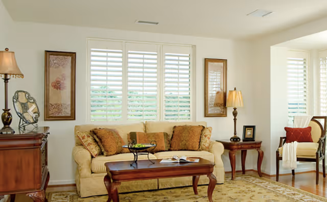 A cozy living room with a beige sofa adorned with patterned cushions, a wooden coffee table with a decorative bowl and an open book, two side tables each with a lamp, framed artwork on the walls, and a window with white plantation shutters letting in natural light.