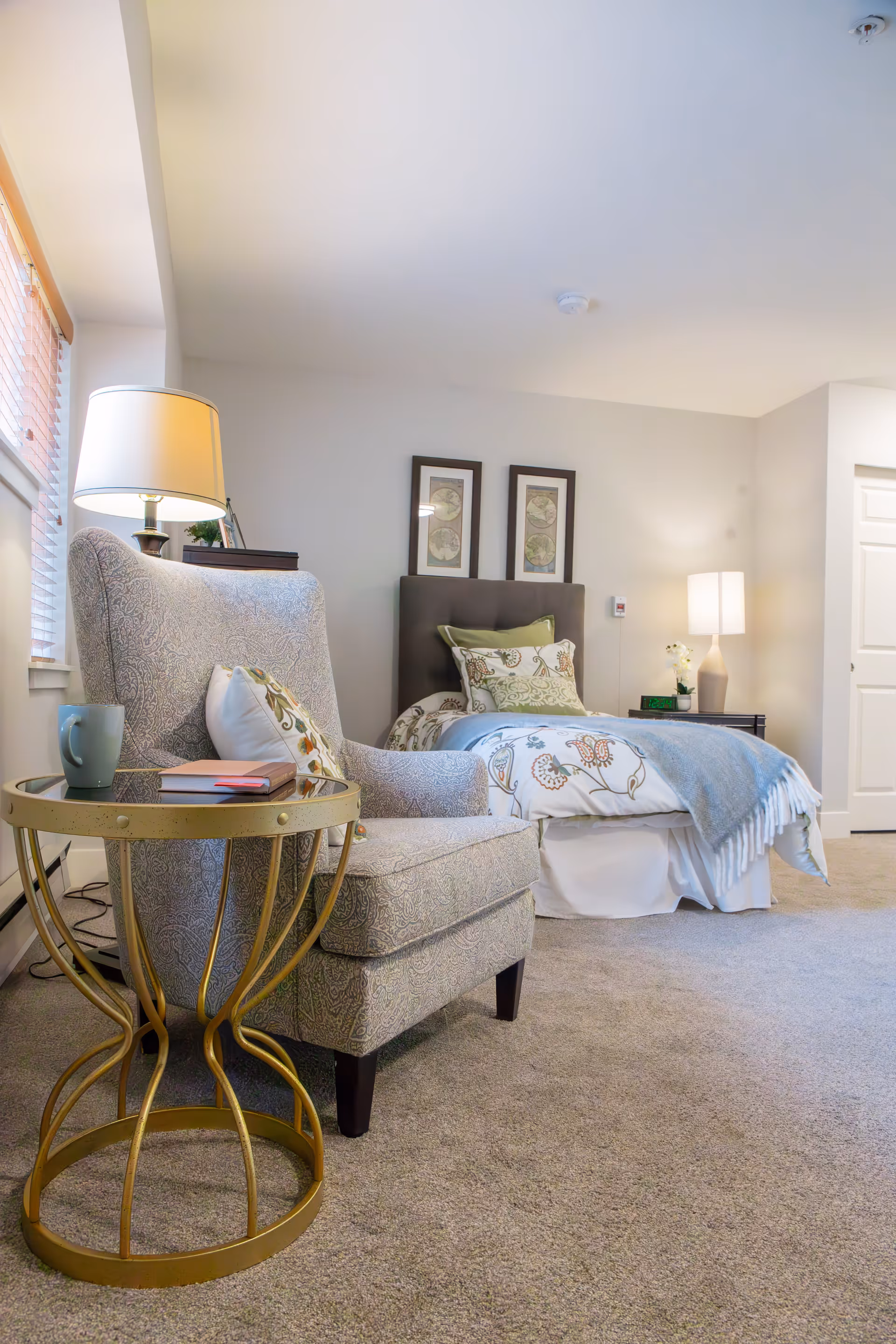 A cozy bedroom featuring a single bed with patterned bedding and multiple pillows, a cushioned armchair with a pillow, a round gold side table holding a mug and a book, two framed artworks on the wall above the bed, and two lamps providing warm lighting.