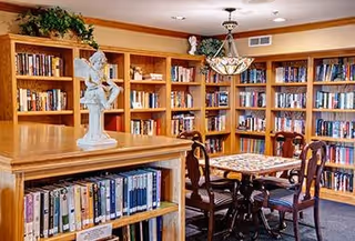 A cozy library room with wooden bookshelves filled with books lining the walls. In the center, there is a wooden table with a decorative chessboard and four wooden chairs with cushioned seats. A decorative statue is placed on a bookshelf in the foreground, and a hanging light fixture is above the table.