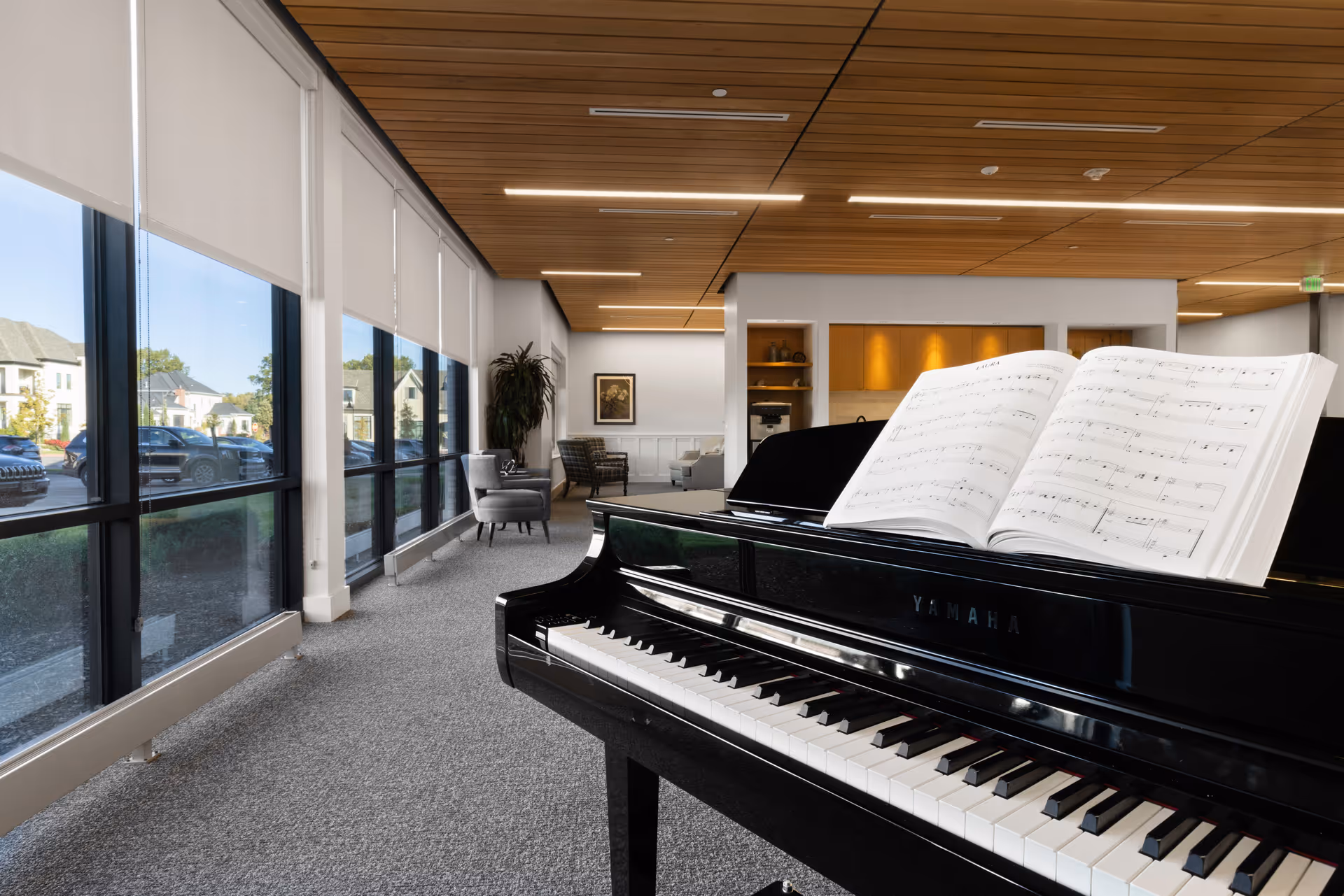 Interior view of a senior living facility lounge area featuring a black Yamaha grand piano with sheet music open on it. The room has large windows with roller shades, gray carpet, modern chairs, a wooden ceiling with recessed lighting, and a small kitchenette area in the background.