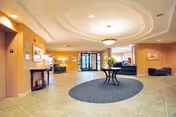 Spacious and well-lit lobby area with beige tiled floors, a round dark rug in the center, a round table with a flower arrangement, seating areas with dark blue sofas, a fireplace on the left wall, and large glass entrance doors at the far end.