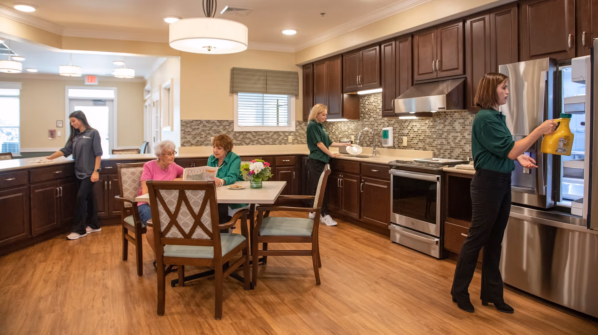 A spacious kitchen and dining area in a senior living facility. Two elderly women are seated at a table with a vase of flowers, one reading a newspaper. Three staff members are engaged in kitchen tasks: one is wiping the counter, another is washing dishes, and the third is taking a jug of orange juice from the refrigerator. The kitchen features wooden cabinets, stainless steel appliances, and a tiled backsplash.