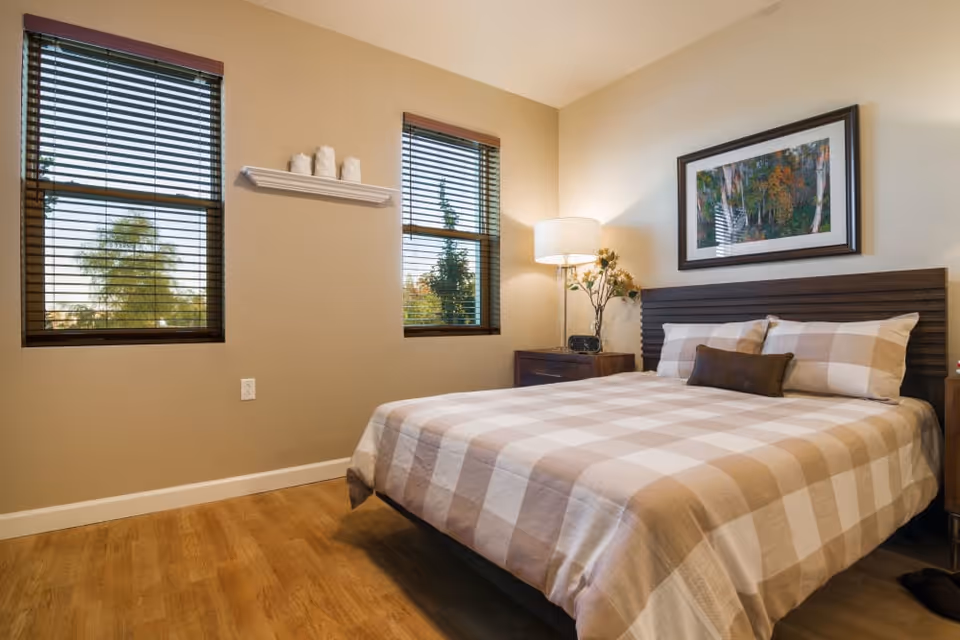 A bedroom with a large bed covered in a beige and white checkered bedspread. The bed has two pillows and a small decorative pillow. There are two windows with wooden blinds on the left wall, a small shelf with three white decorative items between the windows, and a nightstand with a lamp and flowers on the right side of the bed. A framed picture hangs above the headboard. The floor is wooden.