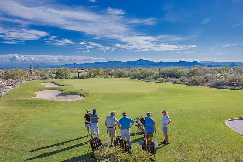 A small group of people standing on a golf green with golf bags, overlooking desert scrub and distant mountains under a blue sky.