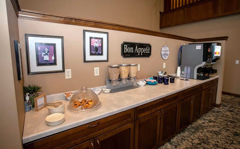 A countertop with breakfast items including cereal dispensers, a covered plate of pastries, bowls, cups, and a beverage dispenser. The wall behind has framed pictures and signs that say 'Bon Appetit' and 'Happiness is a cup of coffee.' The cabinetry below is wooden with multiple drawers and doors.