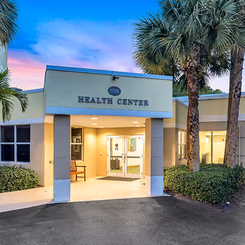 Front entrance of a health center building marked 1725 with lit double doors, a covered entryway, palm trees and landscaping at dusk.