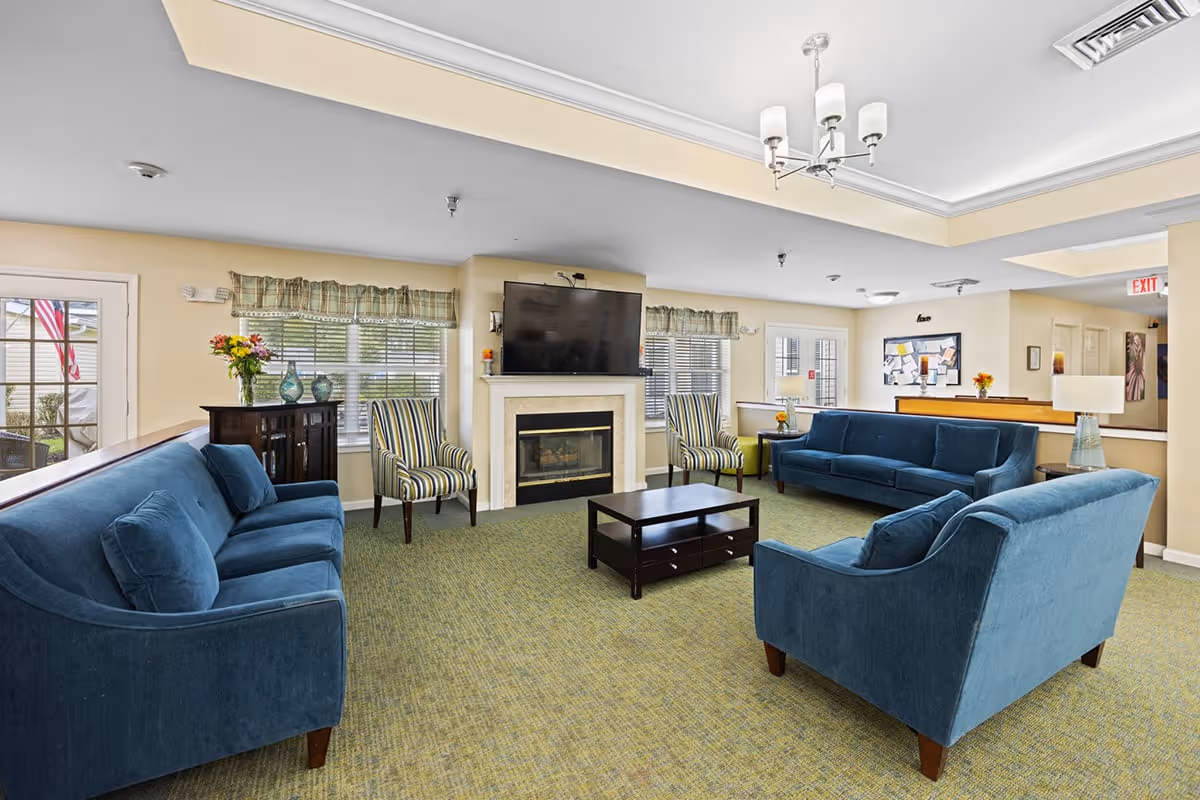 A bright and spacious living room area with blue upholstered sofas and striped armchairs arranged around a dark wooden coffee table. A fireplace with a mounted flat-screen TV above it is centered on one wall, flanked by windows with valance curtains. The room has a green carpet, beige walls, and a ceiling light fixture. There is a door leading outside and a bulletin board on the far wall.
